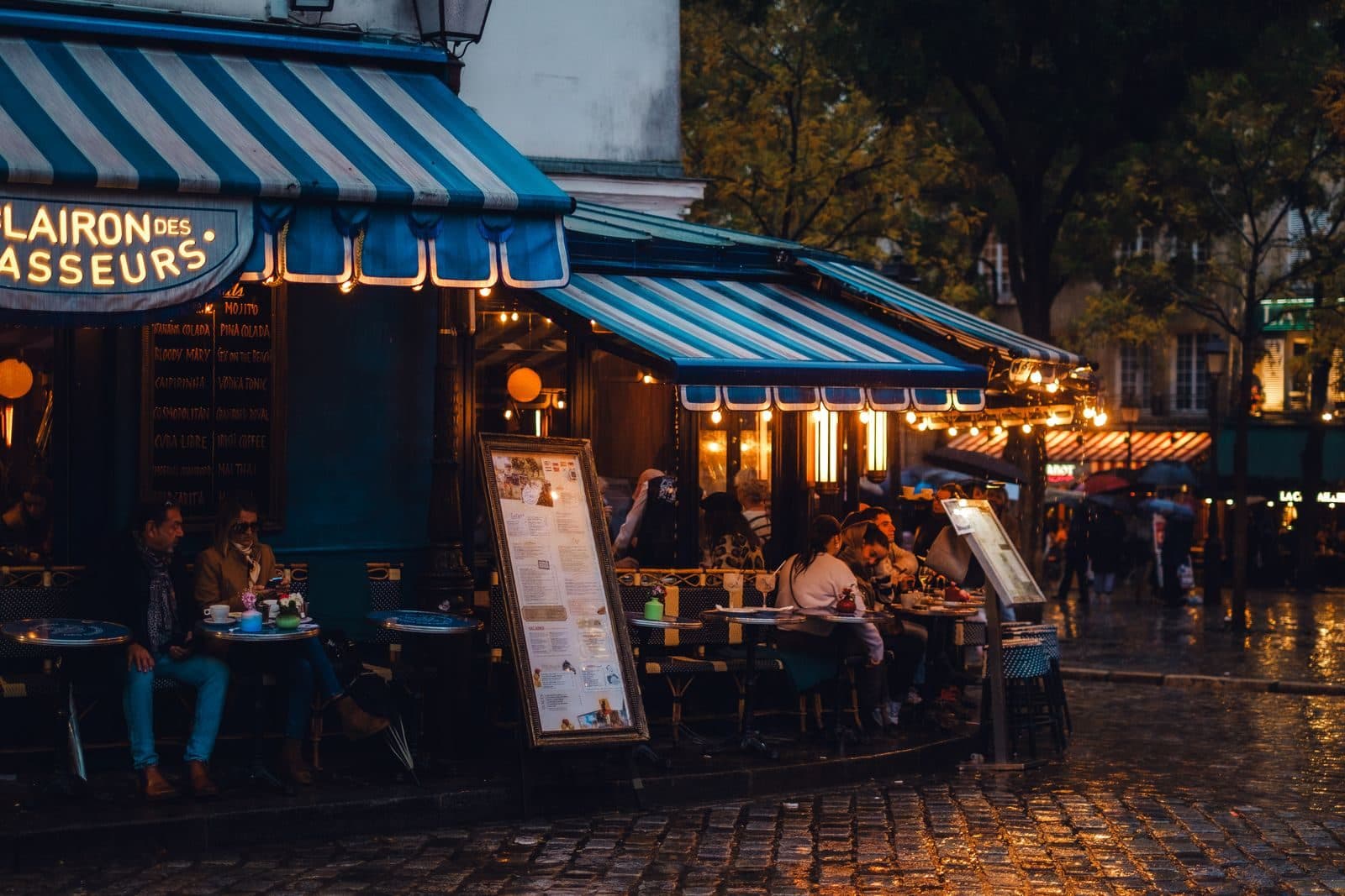 A busy corner café on a rainy evening, its tables full under striped awnings
