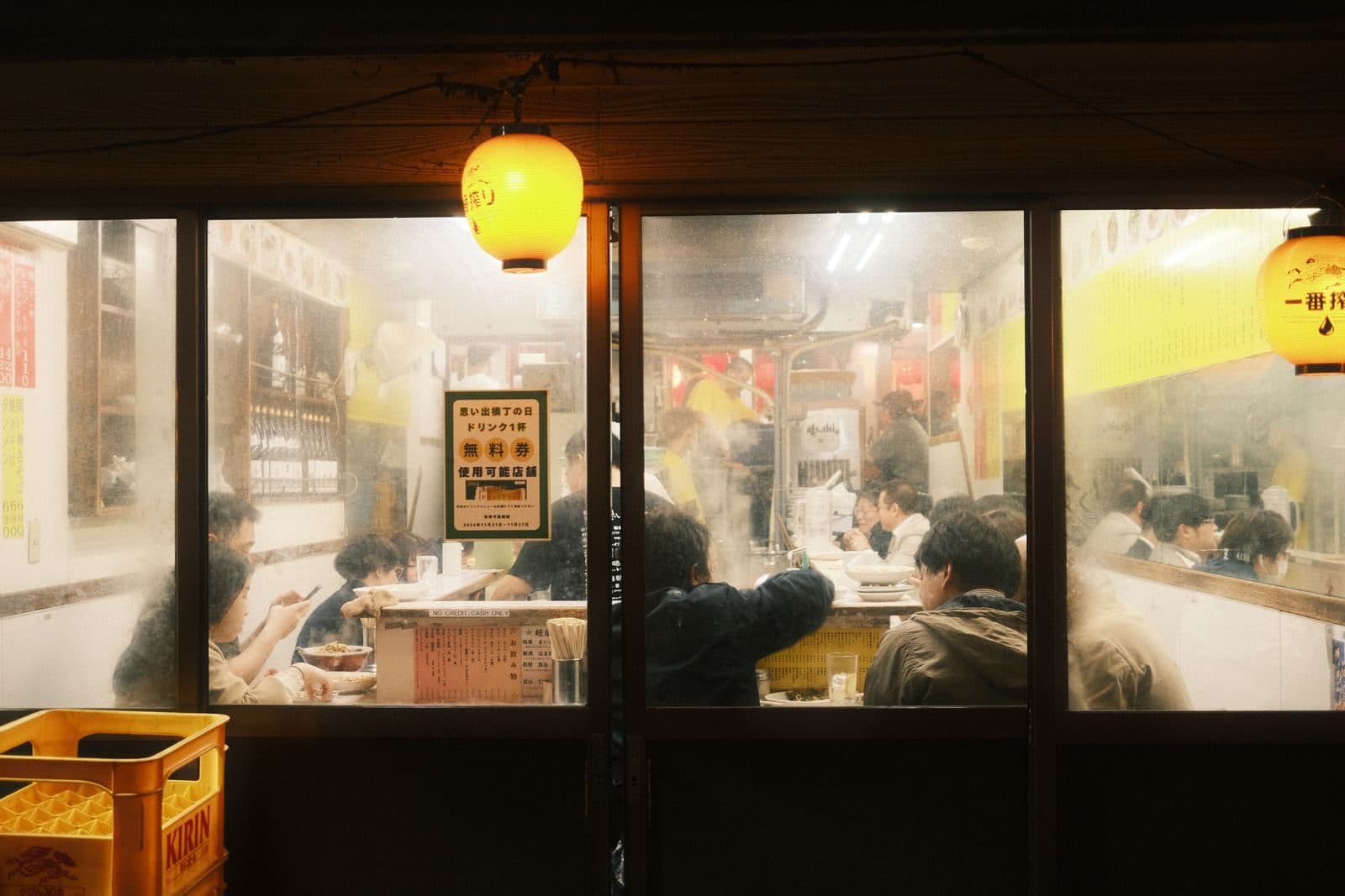 Diners packed into a small restaurant at night, seen through a steamy window under a paper lantern